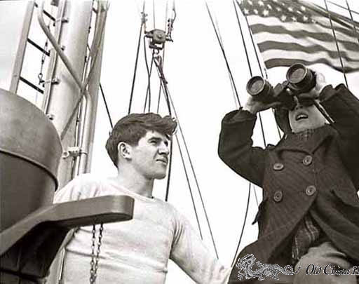 Boy looking through binoculars aboard a ship with man next to boy; US flag in the background