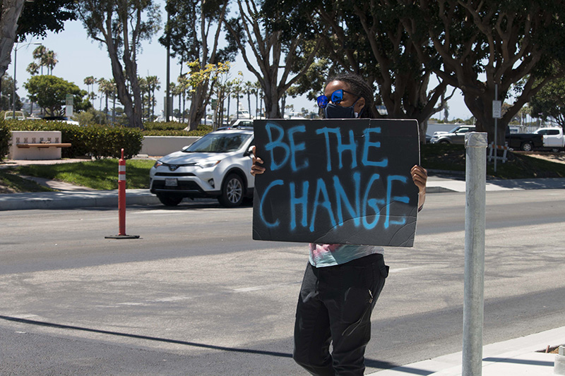 Masked protester holds a handmade sign: BE THE CHANGE