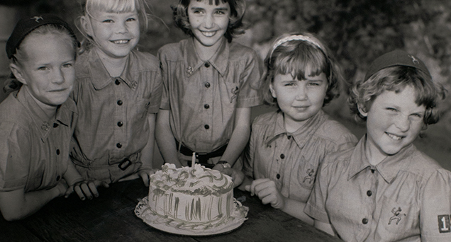 Five young girls around a table looking at the camera with a cake in the center
