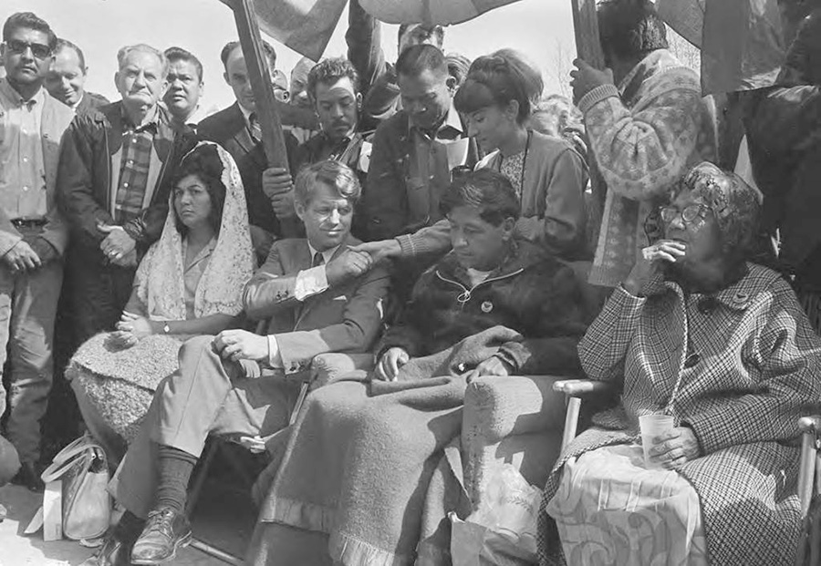 Woman with a white veil, two men, and a woman in a coat sitting in chairs outside, surrounded by a crowd