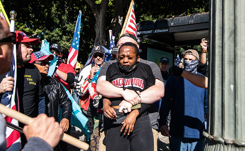 White man holds a black woman with her arms pinned, others with Trump campaign and Stop the Steal gear look on