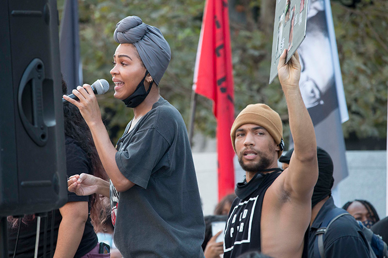 Person standing slightly above the surrounding crowd, speaks though a microphone while a man behind them holds up a sign