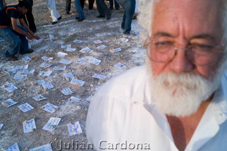 Man with white beard and hair in front of camera with papers scattered on floor in background