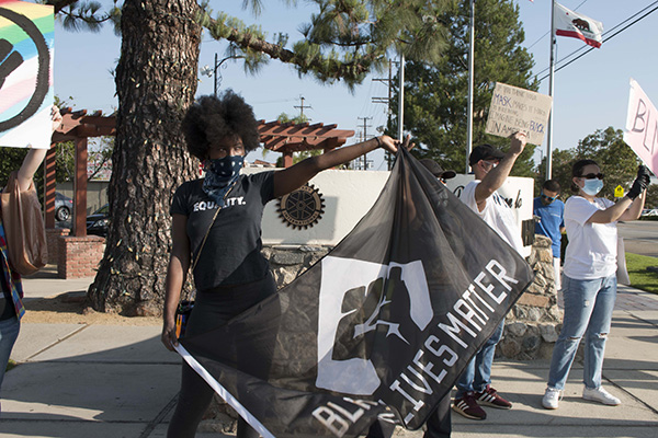 A woman displays a Black Lives Matter flag bearing a raised fist with other protestors holding signs nearby.