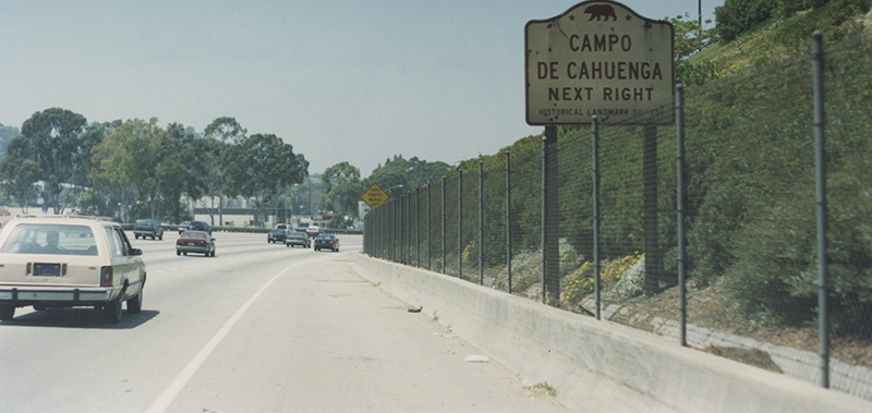 Cars driving along a road, next to road is a sign with a bear icon and the words 'Campo de Cahuenga Next Right'