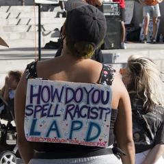 Woman with handmade sign on her back: HOW DO YOU SPELL RACIST? LAPD.  THERE'S BLOOD ON YOUR HANDS PIG