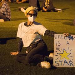 Masked protester seated on the grass, holding a sign: BREONNA IS A STAR