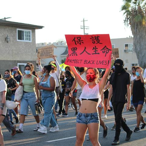 Masked woman marching with others, carrying a sign with English and Chinese: PROTECT BLACK LIVES!