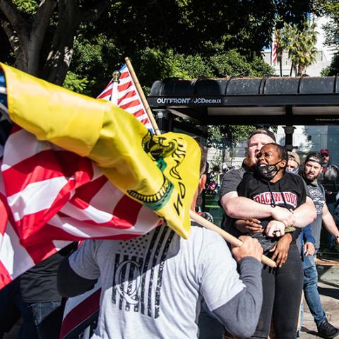 White man holds a black woman with her arms pinned.  Man in a  Qanon shirt, with American and Don't Tread on Me flags in the foreground