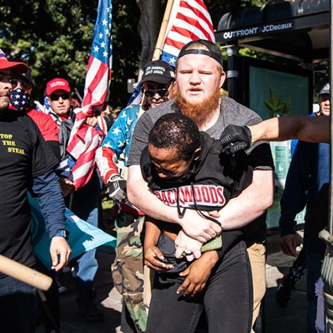 White man holds a black woman with her arms pinned while she is sprayed with mace directly in her eyes, others with Trump campaign and Stop the Steal gear look on