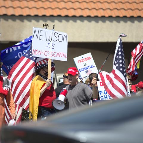 People standing with American flags and signs: NEWSOM IS NON-ESSENTIAL