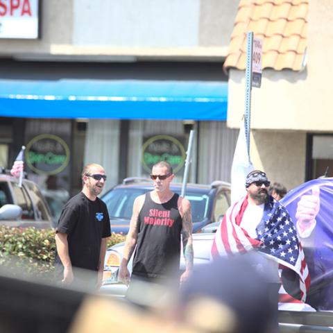 Three men standing on the street, won with a Silent Majority shirt, another with the American flag draped over his shouldersl; Trump banner on the car behind