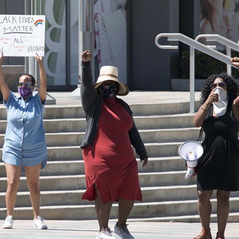 Three women with faces covered stand in protest, two with raised fists and one holding a handmade Black Lives Matter sign