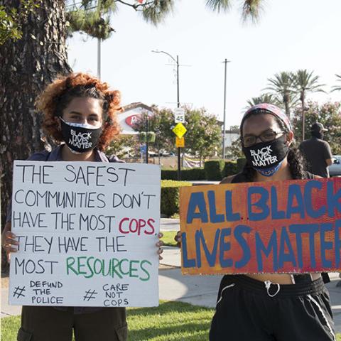  Two women in Black Lives Matter masks holding handmade signs: (1) THE SAFEST COMMUNITIES DON'T HAVE THE MOST COPS.  THEY HAVE THE MOST RESOURCES. #DEFUNDTHEPOLICE #CARENOTCOPS (2) ALL BLACK LIVES MATTER