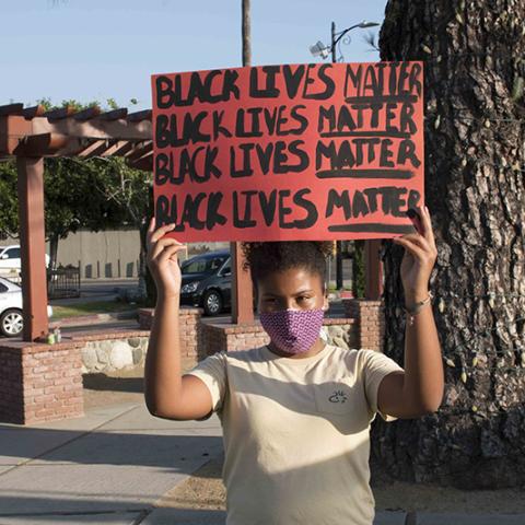 Woman in mask holding a handmade sign with  BLACK LIVES MATTER written four times.