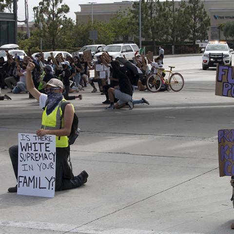 Two masked protesters kneel in the street with handmade signs: (1) IS THERE WHITE SUPREMACY IN YOUR FAMILY? (2) FUCK THE POLICE (3) PROSECUTE KILLER COPS