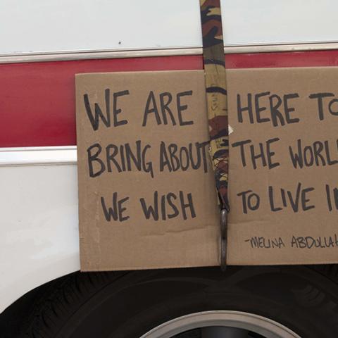 Cardboard sign strapped to a vehicle: WE ARE HERE TO BRING ABOUT THE WORLD WE WISH TO LIVE IN -- MELINA ABDULLAH