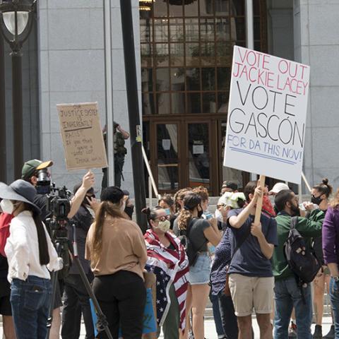 A crowd of masked protesters, with two handmade signs: OUR JUSTICE SYSTEM IS INHERENTLY RACIST.  If you think you're a "GOOD COP" THINK AGAIN; VOTE OUT JACKIE LACEY - VOTE GASCÓN FOR DA THIS NOV!