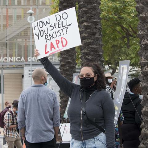 Masked woman holding handmade sign: HOW DO YOU SPELL RACIST? LAPD