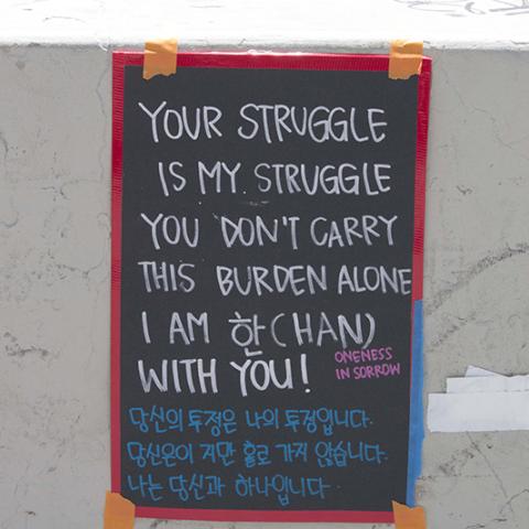 Sign in both English and Korean languages taped to a wall: YOUR STRUGGLE IS MY STRUGGLE.  YOU DON'T CARRY THIS BURDEN ALONE. I AM WITH YOU