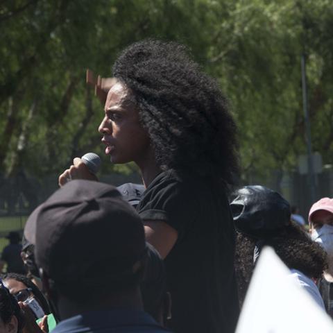 Person standing slightly above the surrounding crowd, speaks though a microphone