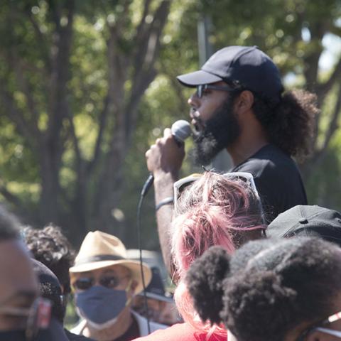 Man standing slightly above the surrounding crowd, speaks though a microphone
