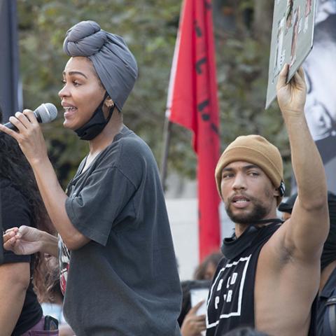 Person standing slightly above the surrounding crowd, speaks though a microphone while a man behind them holds up a sign