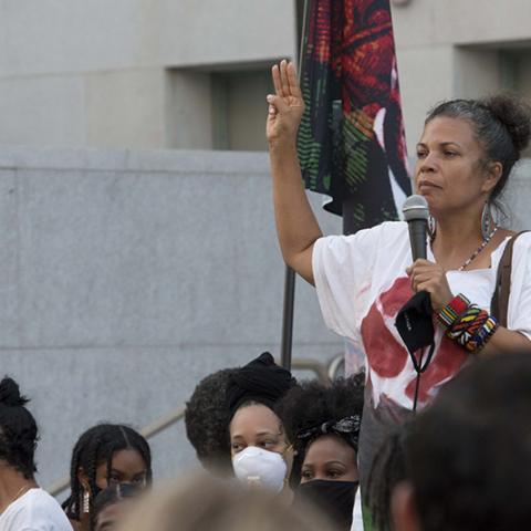 Woman holds up 3 fingers in right hand, microphone in left hand.