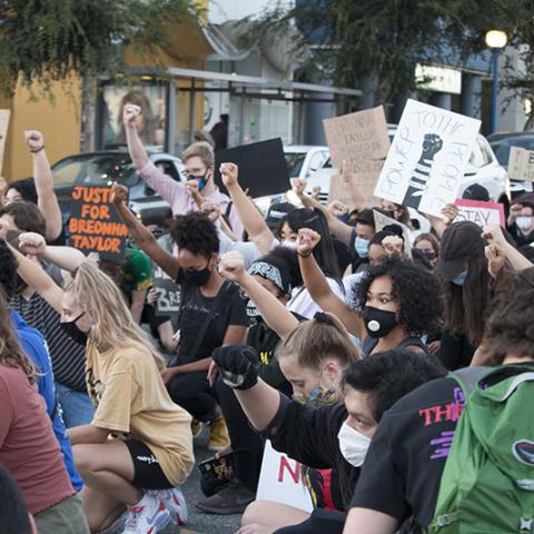 Large group of masked people kneeling with fists raised and handmade signs: JUSTICE FOR BREONNA TAYLOR; BREONNA TAYLOR SHOULD BE HERE; POWER TO THE PEOPLE; BLACK LIVES MATTER