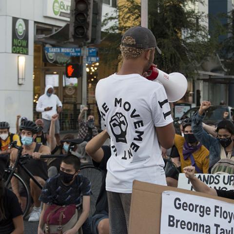 A speaker with a megaphone addresses a crowd kneeling in the street