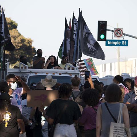 Group of masked people surrounding a vehicle carrying multiple flags include BLACK LIVES MATTER and a photo of Malcolm X