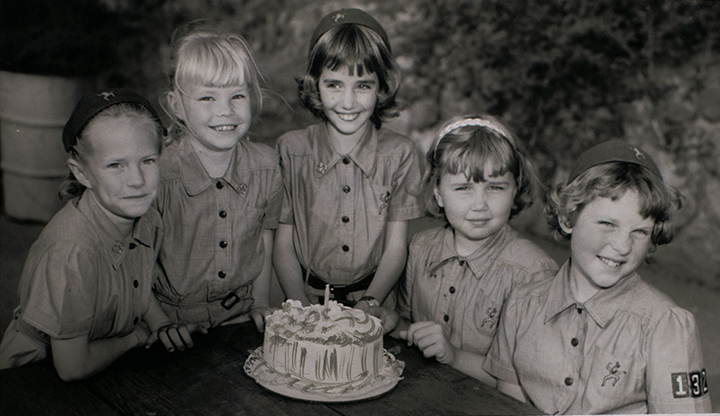 Five young girls around a table looking at the camera with a cake in the center