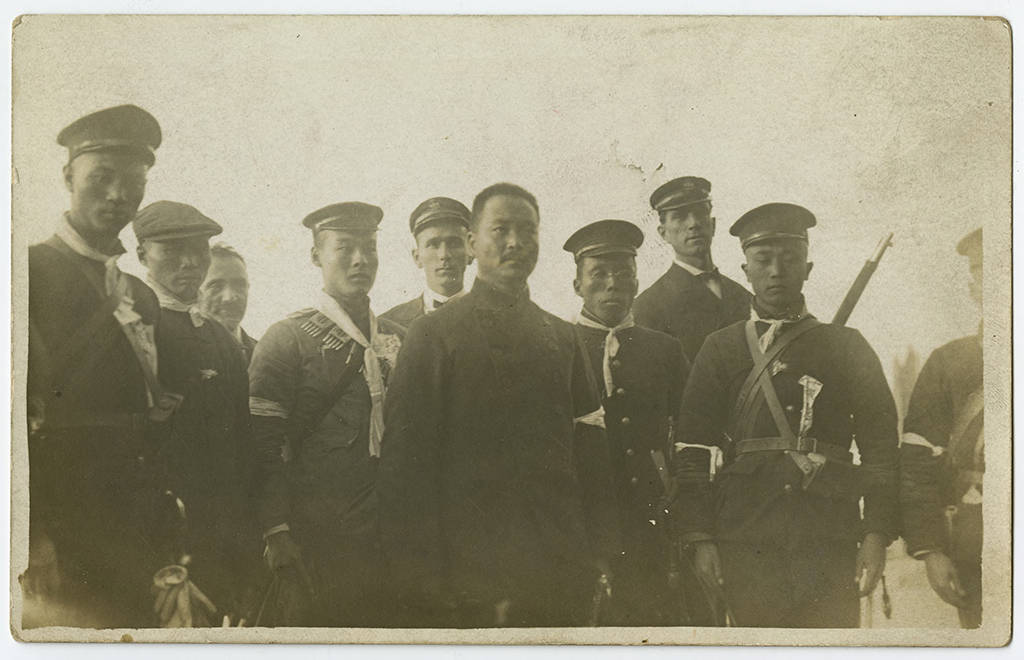 Armed Chinese or Japanese soldiers in uniform stand in the foreground and three American, British, or European soldiers stand in the background
