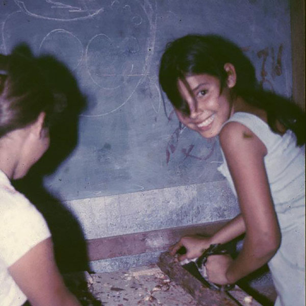 Child looking at camera while carving a piece of wood; a woman is working next to her.