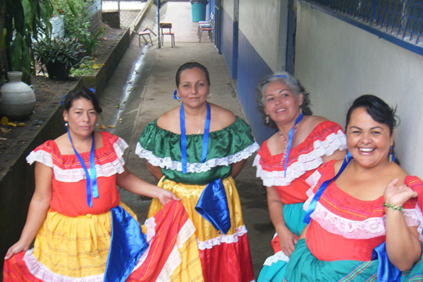 Group of women in folkloric costumes posing in a corridor outside a building.