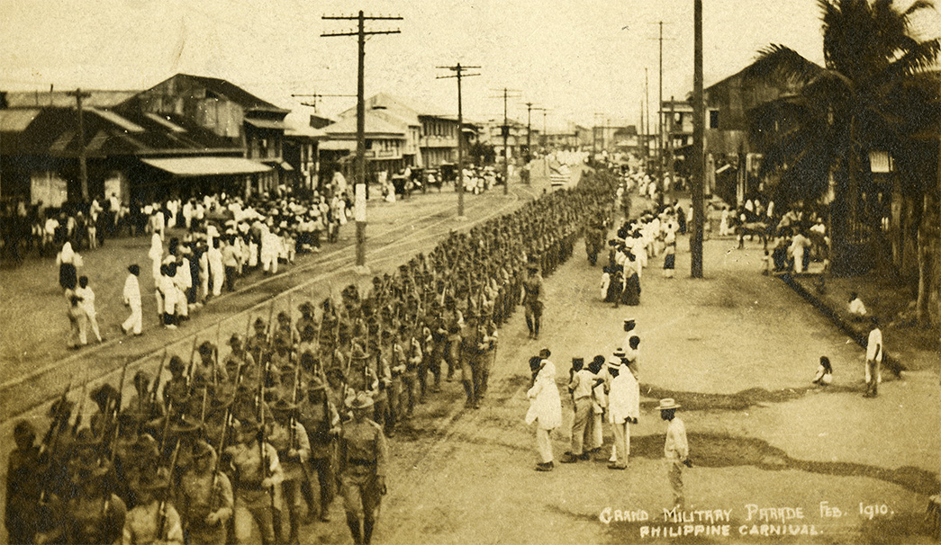 Soldiers in uniform carrying rifles march down a wide boulevard as civilians look on. Buildings and powerlines are in the background.