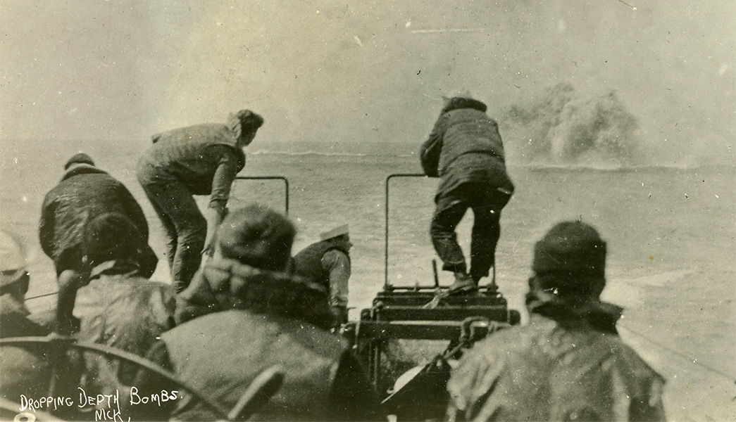 Photograph of sailors watching in the forground as a bomb explodes underwater in the distance.