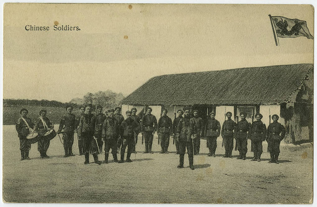 Chinese soldiers with rifles and bayonets for a semicircle infront of a single story building with a thatched roof. Two soldiers at the right of the semi circle carry drums.
