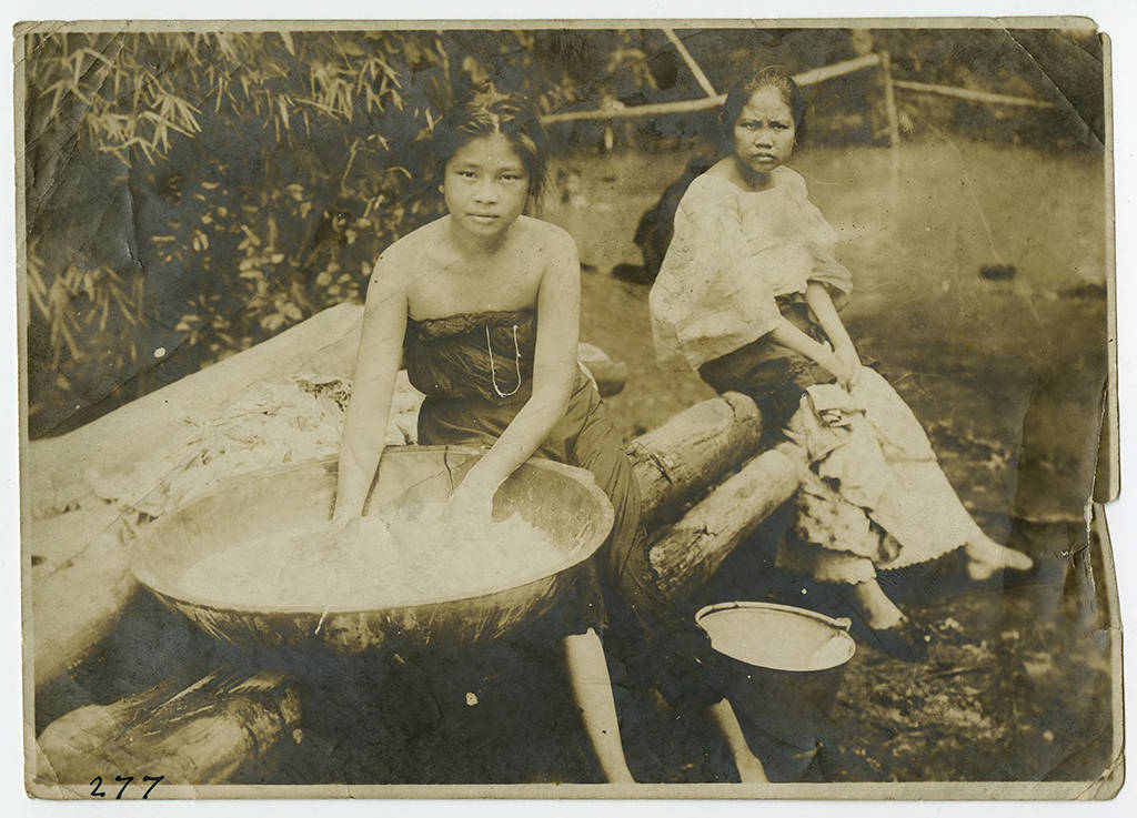 Portrait of two young women sitting on logs washing clothing in a large bowl.